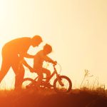 Silhouette of father teaching little daughter to ride a bike at meadow during sunset. Caring father teaches her daughter to ride bicycle
