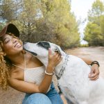 young pretty girl with curly hair and hat petting her dog outdoors in the hills in sunlight laughing and bonding. Dog licking her chick.